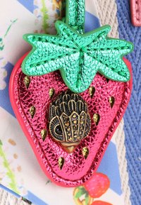 Accessorio a forma di fragola di un vivace colore rosa, con una finitura texture, foglie metalliche verdi e dettagli dorati, caratterizzato da un elemento decorativo a forma di uccello nero.