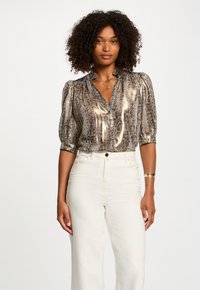 Blouse en bronze métallique avec un motif serpent, devant à boutons et manches bouffantes, associée à un pantalon large crème.