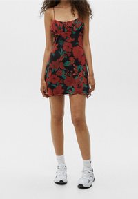 Robe florale avec des motifs rouges et verts sur un fond noir, dotée de fines bretelles et d'un col volanté, avec une texture douce et légère.