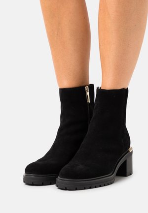 Sorte ruskinds ankelstøvler med chunky hæle og lynlåse i siden, båret på bar hud mod en hvid baggrund.