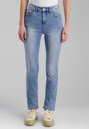 Person trägt hellblaue Jeans mit geradem Bein und cremefarbene Schnürsneaker, steht vor einem schlichten grauen Hintergrund.