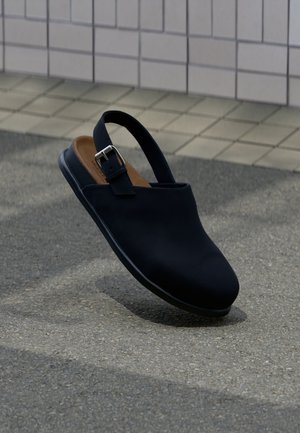 Μαύρο κλειστό παπούτσι slingback με ασημένιο λουράκι αγκράφας, καφέ πάτος, αιωρείται πάνω από γκρίζο άσφαλτο με φόντο πλακιδωτό τοίχο.