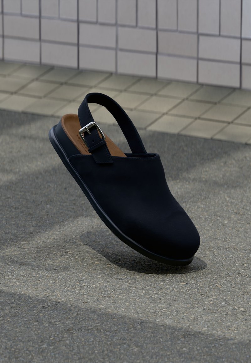 Μαύρο κλειστό παπούτσι slingback με ασημένιο λουράκι αγκράφας, καφέ πάτος, αιωρείται πάνω από γκρίζο άσφαλτο με φόντο πλακιδωτό τοίχο.