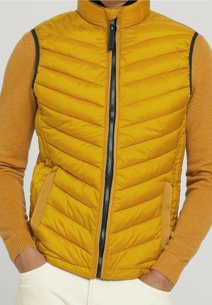 Żółta pikowana bezrękawnik z zamkiem błyskawicznym z przodu, noszona na teksturowanym swetrze z długim rękawem, ręce w kieszeniach, na neutralnym tle.