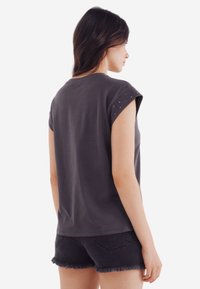 Grå kortærmet t-shirt med opkrøllede ærmer, der har en glat tekstur og små dekorative detaljer på skuldrene. Parret med sorte shorts.