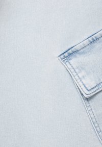 Tissu en denim bleu clair avec une texture subtile, comprenant un détail de poche pliée et des surpiqûres contrastantes le long des bords.