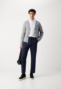 Grå ull cardigan over en hvit skjorte med knapper, kombinert med marineblå bukser og svarte joggesko. Modellen holder en svart skulderveske.