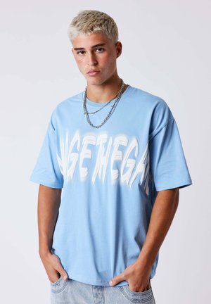 Jeune homme aux cheveux courts décolorés porte un t-shirt oversize bleu clair avec un grand texte blanc et des chaînes argentées superposées, mains dans les poches.