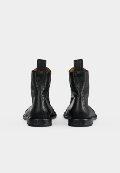 Ζευγάρι μαύρα δερμάτινα μποτάκια αστραγάλου με ταινίες για τράβηγμα και διακριτικές λεπτομέρειες brogue, από πίσω σε λευκό φόντο.