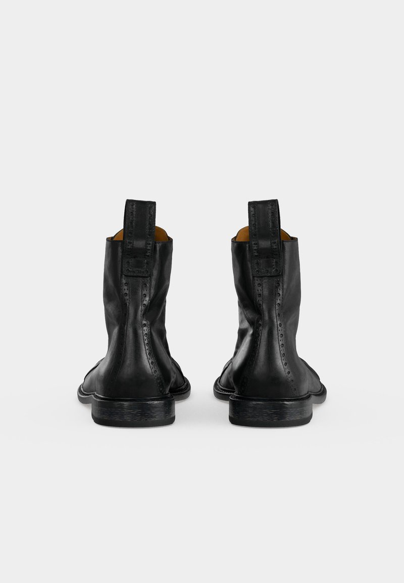 Paar schwarze Leder-Stiefeletten mit Zuglaschen und subtiler Brogue-Verzierung, von hinten auf weißem Hintergrund betrachtet.