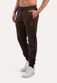 Brune sweatpants med elastisk talje og snøre, sidelommer og et lille logo mærke, parret med hvide sneakers.