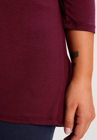 Langärmliges, burgunderfarbenes Oberteil mit glatter Textur und einem abgerundeten Saum. Das Shirt sitzt locker am Oberkörper und hat einen seitlichen Schlitz.