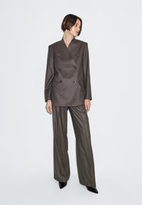 Blazer marrone doppiopetto con spalle marcate, abbinato a pantaloni a gamba larga. Entrambi realizzati in tessuto testurizzato. Presenta bottoni coordinati.