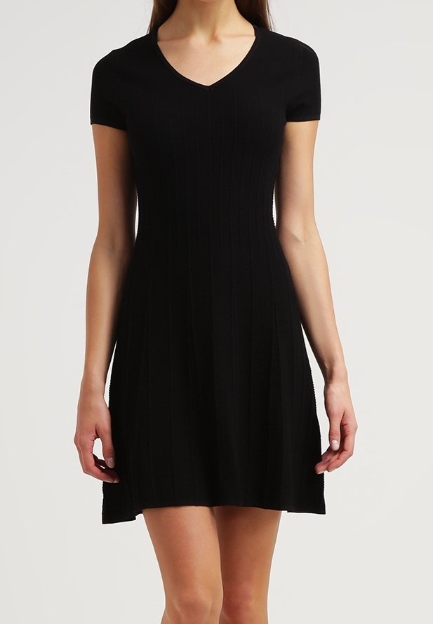 Femme portant une robe noire courte à manches courtes et col en V, debout devant un fond clair uni.