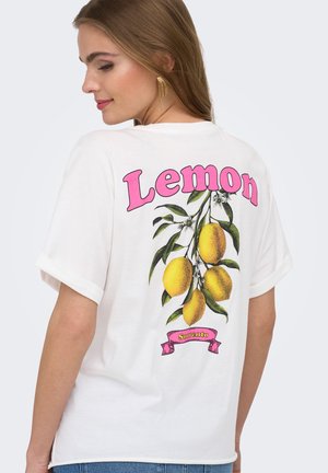 Młoda kobieta z długimi brązowymi włosami nosząca białą koszulkę z różowym napisem "Lemon" i ilustracją cytryn oraz liści na plecach.