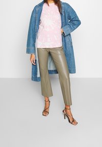 Kvinna iklädd en rosa tie-dye tröja, blå denimjacka, olivgröna läderbyxor och svarta sandaletter med hög klack, i stående pose.