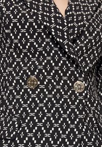 Schwarz-weißer Blazer mit geometrischem Muster, breiten Revers, doppelten goldenen Knöpfen und Klapptaschen.