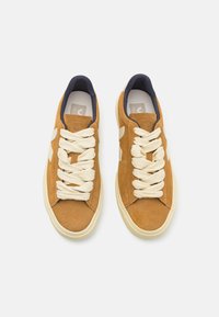 Tan semskinnesko med kremfargede lisser, marineblå foring og beige gummisåle. Har et enkelt design med en minimal logo på siden.