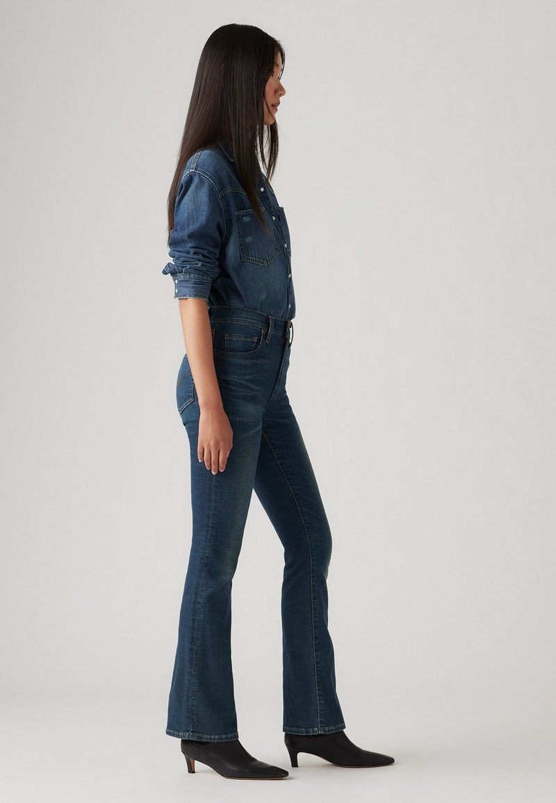Denimhemd mit Knopfleiste, spitzen Kragen und zwei Brusttaschen, kombiniert mit dunkelblauen Schlagjeans und schwarzen angeknöpften Stiefeletten.