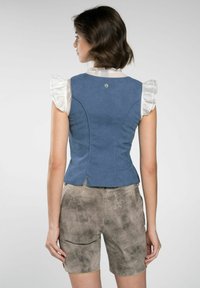Blauwe denimvest met een getailleerde pasvorm, voorzien van ruches mouwen in het wit, gestikte details en een klein logo op de achterkant. Gecombineerd met beige shorts.
