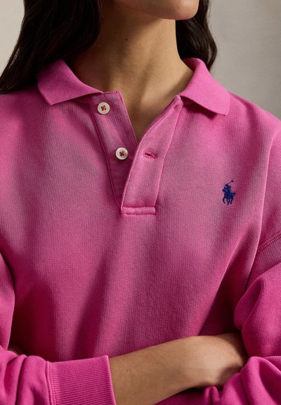 Ροζ πόλο μπλούζα από απαλό βαμβάκι με γιακά, εμπρόσθιο άνοιγμα με δύο κουμπιά και κεντημένο λογότυπο σε ναυτικό μπλε στο αριστερό μέρος του στήθους.