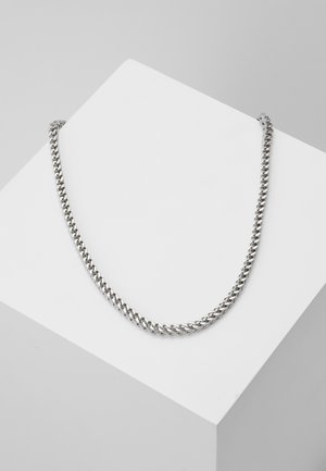 Collier en chaîne argentée avec des maillons entrelacés, design plat, finition réfléchissante, reposant sur une surface d'exposition géométrique blanche.