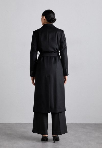 Culoarea neagră a paltonului lung prezintă un design ajustat, cu o curea la talie și mâneci largi, fiind asortat cu o pereche de pantaloni largi și pantofi cu toc înalt.