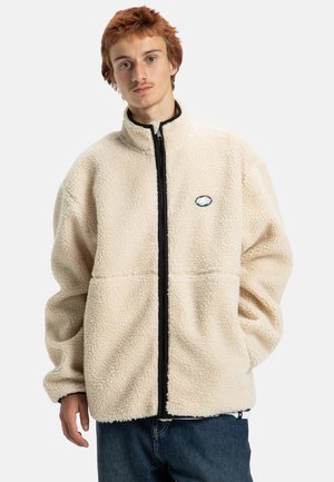 Chaqueta de forro polar color crema con cuello alto, cremallera frontal completa y detalles en negro. Presenta un parche con el logo en el lado izquierdo del pecho y un tejido texturizado.