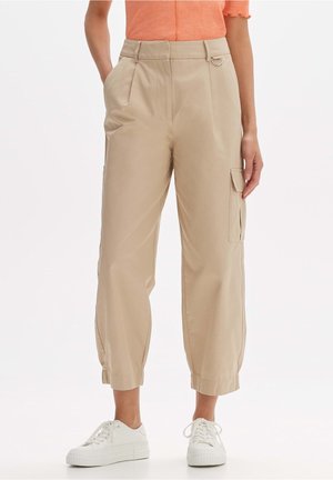 Beige korte cargobukser med sidelommer, elastiske manchetter og en lille metalringdetalje, båret med hvide sneakers og en orange top.