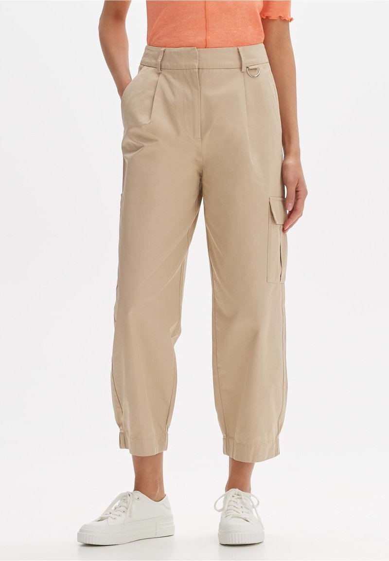Pantalon cargo beige court avec poches latérales, poignets élastiques et un petit détail anneau en métal, porté avec des baskets blanches et un haut orange.