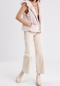 Fuskepelsvest i blød beige med hætte, der har trykknapper, sidelommer og en struktureret overflade, kombineret med lyse cremefarvede bukser og beige støvler.