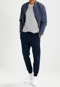 Navy bomber jacket over white and navy striped shirt, paired with navy joggers and white sneakers. Smooth fabric and elastic cuffs.