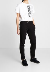 Weißes, kurzes T-Shirt mit schwarzem "PUMA"-Logo an der Seite, kombiniert mit schwarzen Jogginghosen mit Kordelzug und Taschen.