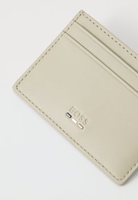 Portafoglio in pelle di colore beige chiaro. Presenta quattro scomparti per carte, cuciture a vista e logo impressa con accenti neri, bianchi e arancioni.