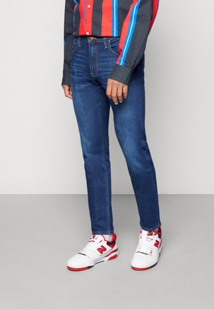 Jean en denim bleu avec une coupe droite, présentant un design classique à cinq poches. Associé à une chemise à rayures multicolores et des baskets blanches.