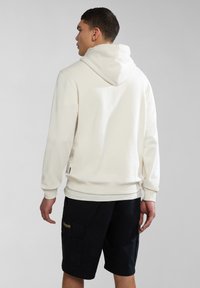Sudadera color crema con capucha ajustable, puños de canalé y dobladillo. Combinada con pantalones cortos oscuros que tienen bolsillos laterales y un parche de marca.