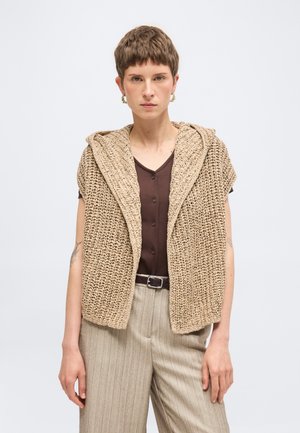 Osoba ubrana w krótki brązowy koszulę na guziki, beżową dzianą kamizelkę bez rękawów z kapturem oraz beżowe spodnie w cienkie prążki z czarnym paskiem.