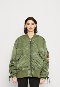 Olive green bomber jacket with a zip front, ribbed cuffs, and oversized fit. Features side pockets and a decorative strap on one sleeve.