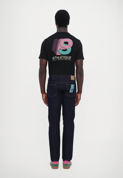 Camiseta negra con gráfico colorido, combinada con jeans de mezclilla oscuros con costuras naranjas y una etiqueta azul claro en el bolsillo trasero. Zapatillas multicolores.