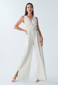 Lyse beige jumpsuit med V-hals, omslagsdesign, indsnøret talje og vide ben med sideslids. Fremhævet med en snøre.