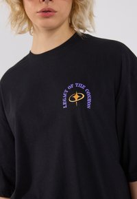 Black cotton t-shirt featuring a front graphic with the text "LEGACY OF THE COSMOS" in purple and gold, short sleeves, and a relaxed fit.