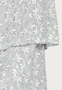 Szary przezroczysty materiał z misternym białym haftem kwiatowym, ozdobiony cekinami i koralikami, z falbaniastym dołem.