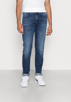 Hombre con vaqueros azules de corte slim remangados y zapatillas blancas, de pie frente a un fondo claro y liso.