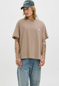 Brun bomulds T-shirt med korte ærmer og løs pasform, der har et lille broderet logo på brystet og bæres med blå denim jeans.