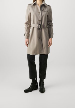 Frau trägt einen beigen Trenchcoat mit Gürtel, schwarze kurze Hosen und schwarze Stiefeletten, sie steht vor einem schlichten hellen Hintergrund.