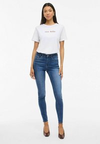 T-shirt blanche avec le texte "ciao bella" en rouge, associée à un jean slim bleu et à des chaussures à talons marron. Design simple, style décontracté.