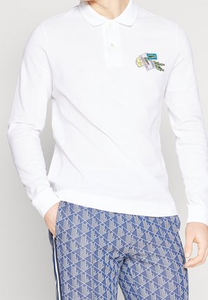 Polo blanc à manches longues avec un petit écusson coloré sur la poitrine, associé à un pantalon bleu à motifs avec formes géométriques blanches et bandes sur les côtés.