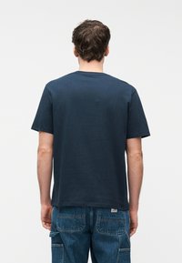 Άνδρας που στέκεται με την πλάτη στραμμένη, φορώντας μονόχρωμο μπλε σκούρο κοντομάνικο μπλουζάκι και μπλε τζιν παντελόνι cargo, μπροστά σε λευκό φόντο.