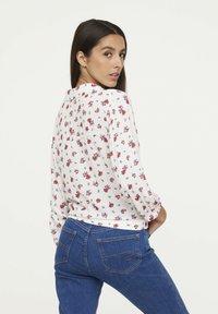 Langarm-Bluse in Weiß mit rotem und blauem Blumenmuster, kombiniert mit enganliegenden blauen Jeans aus Denim, die zwei Gesäßtaschen und gelbe Nähte aufweisen.