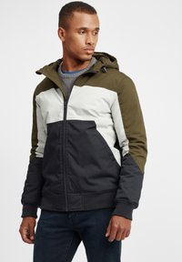 Hooded jacket featuring color blocks in olive green, black, and cream, with a zip front, elastic cuffs, and a textured interior.
